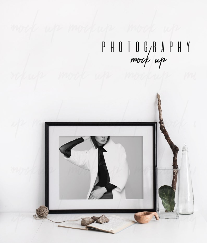 May include: Black and white photo of a woman wearing a white coat and black top in a black frame on a white table with a vase of greenery and a wooden bowl.