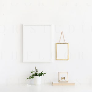 May include: A white wall with two picture frames, one white and one gold, hanging on the wall. A small potted plant sits on a white surface in front of the wall. A gold geometric candle holder sits on a book on the surface.