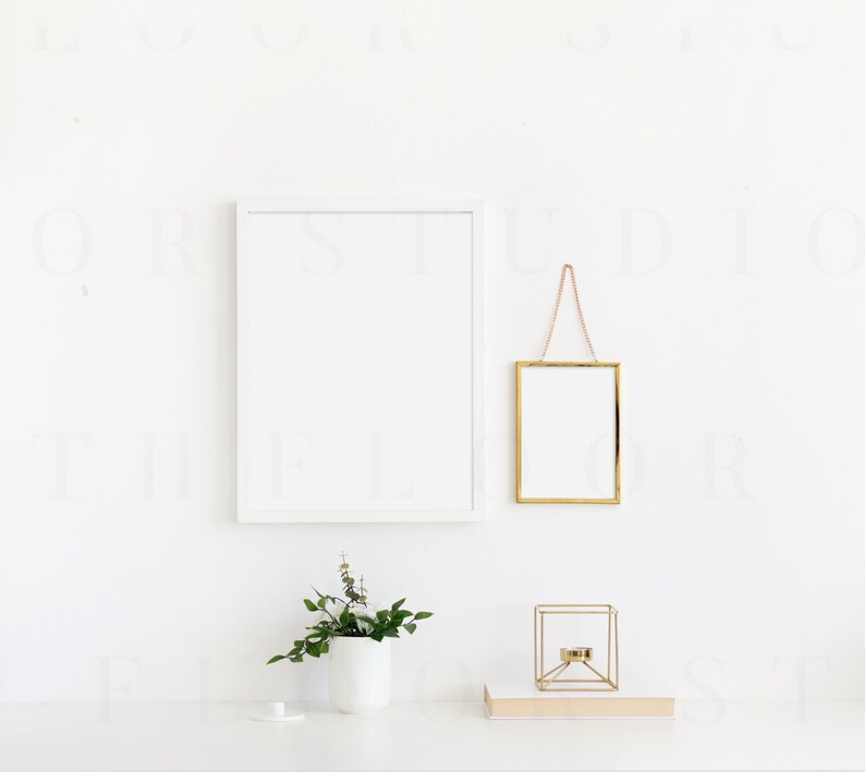 May include: A white wall with two picture frames, one white and one gold, hanging on the wall. A small potted plant sits on a white surface in front of the wall. A gold geometric candle holder sits on a book on the surface.