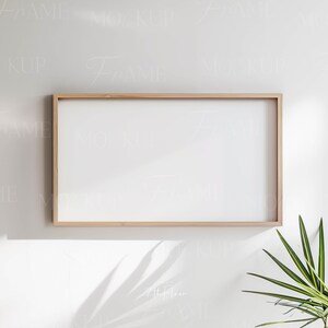 May include: A light wood picture frame with a white mat and a white background. The frame is hanging on a white wall with a green plant in the corner.
