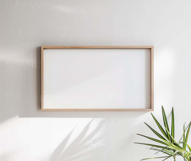 May include: A light wood picture frame with a white mat and a white background. The frame is hanging on a white wall with a green plant in the corner.