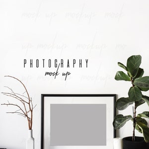 May include: A black picture frame with a white mat and a gray background sits on a white shelf with a white vase, a brown twig, a ball of twine, and a white candle next to a potted plant with green leaves. The text "PHOTOGRAPHY mock up" is written in black on the white wall behind the frame.