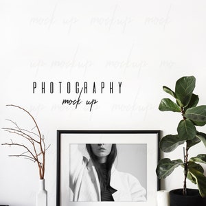 May include: Black and white photo of a person wearing a white jacket and black turtleneck in a black frame on a white shelf with a white vase and a branch with brown twigs, a white candle and a potted plant with green leaves in a black pot.