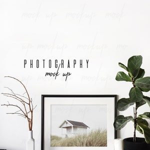 May include: A black framed photo of a white house with a black roof on a grassy hill. The photo is on a white shelf with a white vase with brown branches, a ball of twine, and a white candle. A potted plant with green leaves is on the right side of the shelf. The text "PHOTOGRAPHY mock up" is written in black on the white wall behind the shelf.