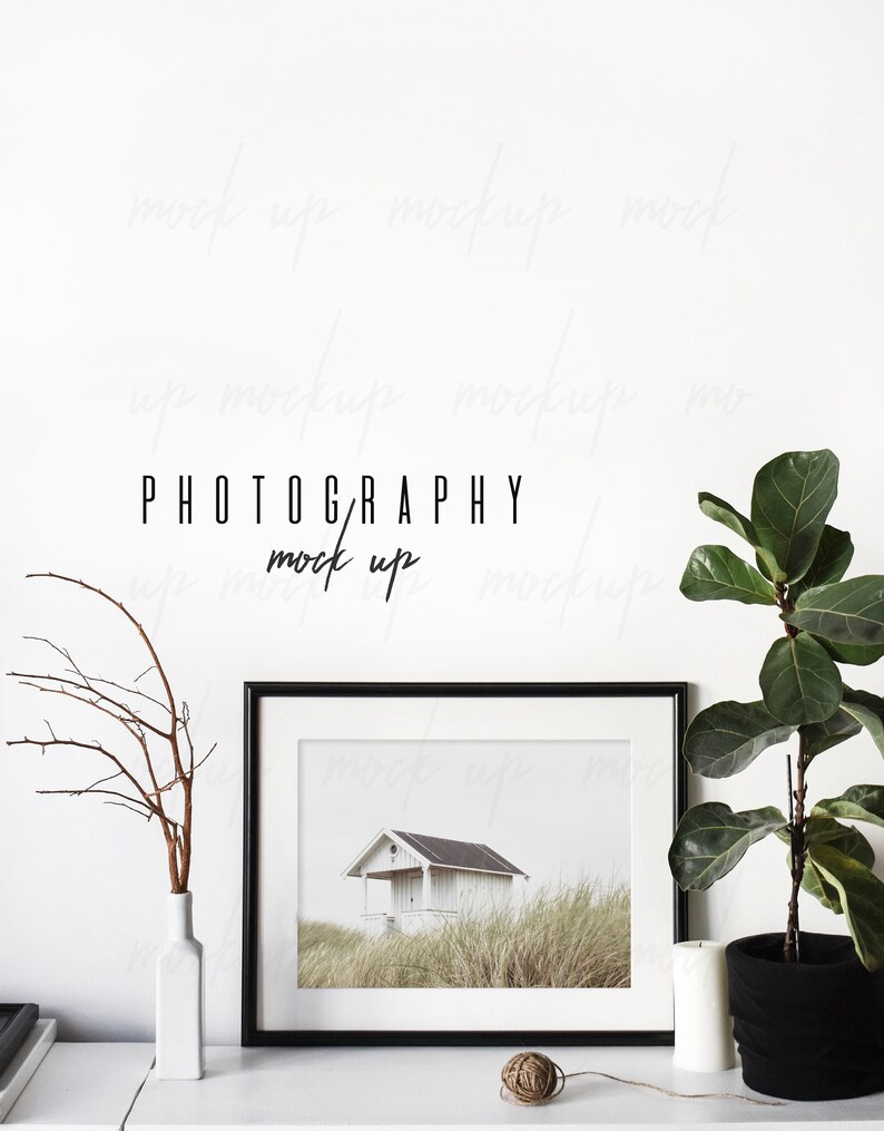 May include: A black framed photo of a white house with a black roof on a grassy hill. The photo is on a white shelf with a white vase with brown branches, a ball of twine, and a white candle. A potted plant with green leaves is on the right side of the shelf. The text "PHOTOGRAPHY mock up" is written in black on the white wall behind the shelf.
