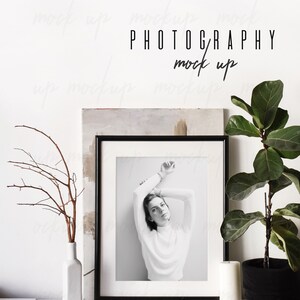 May include: A black framed photo of a woman in a white turtleneck sweater with her arms raised above her head. The photo is on a white shelf with a vase of dried branches, a candle, a ball of twine, and a potted plant with green leaves.