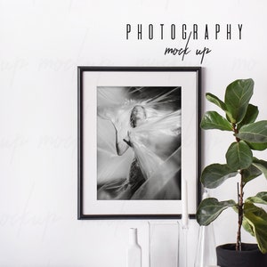 May include: Black and white photograph of a person in a plastic sheet, framed in a black frame, on a white wall with a plant and decorative items on a white shelf below. The text "PHOTOGRAPHY mock up" is above the frame.