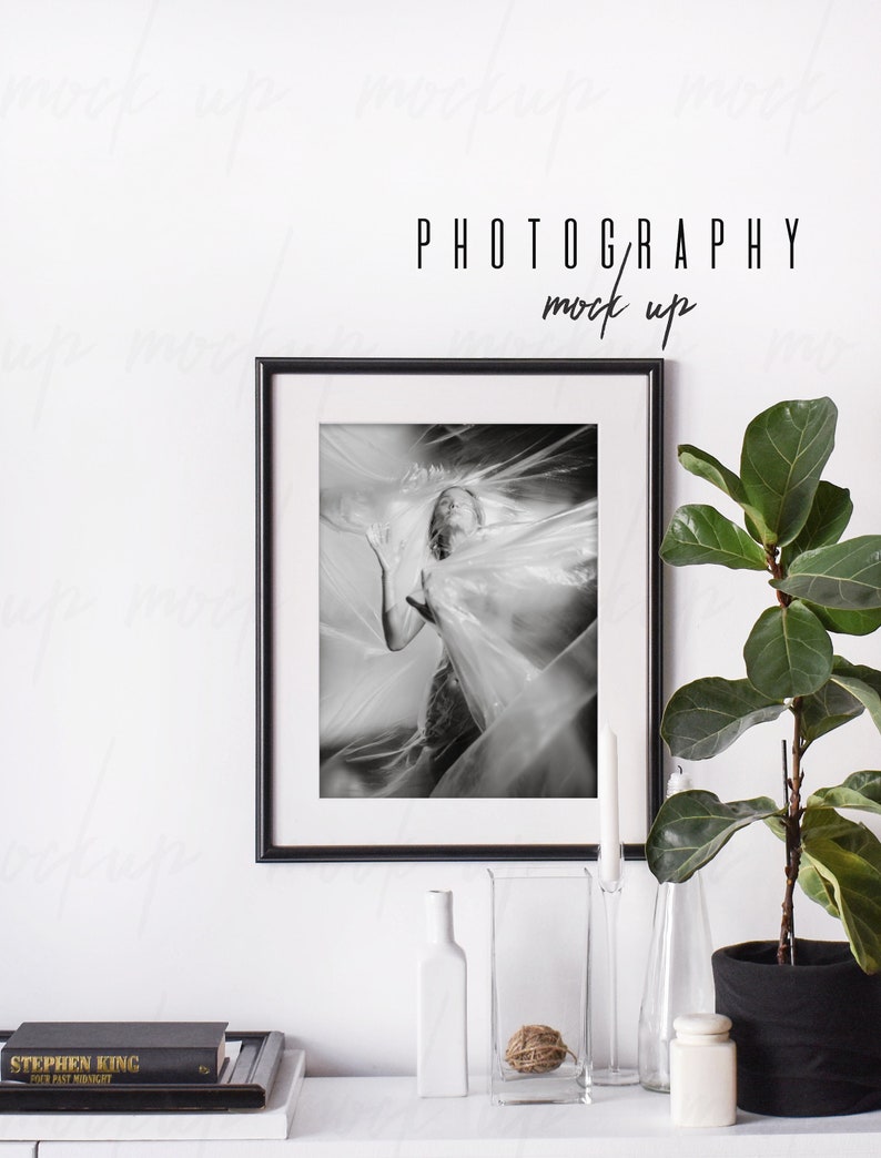 May include: Black and white photograph of a person in a plastic sheet, framed in a black frame, on a white wall with a plant and decorative items on a white shelf below. The text "PHOTOGRAPHY mock up" is above the frame.