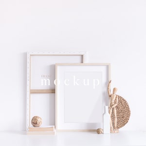 May include: Two wooden picture frames with white mounts, one larger than the other, are leaning against a white wall. The frames are empty and ready for a photo or artwork. A wooden figurine, a small ball, and a white vase are on a white surface in front of the frames.