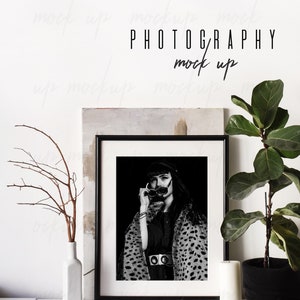 May include: Black and white photo of a woman wearing a leopard print coat and sunglasses in a black frame. The photo is framed in a black frame and is displayed on a white shelf with a vase, a potted plant, and a ball of twine.