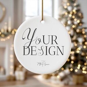 May include: A white, round ceramic ornament with the words "Your Design" in black script and block letters. The ornament is suspended by a gold cord. The text "7th Floor Studio" is below the main text. A Christmas tree is in the background.