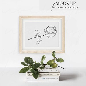May include: A wooden frame with a white mat and a black line drawing of a single peony flower. The frame is sitting on a stack of three books with French titles. A green leafy branch is in front of the frame.