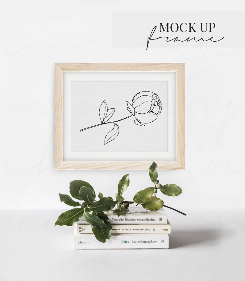 May include: A wooden frame with a white mat and a black line drawing of a single peony flower. The frame is sitting on a stack of three books with French titles. A green leafy branch is in front of the frame.