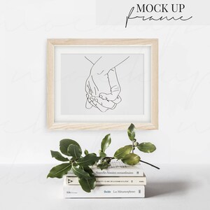 May include: A wooden frame with a white mat and a black line drawing of two hands holding each other. The frame is sitting on a stack of three books with the titles "Nouvelles histoires extraordinaires", "MAZETIC DE LA TOME DA COM", and "Ovide. Les M&eacute;tamorphoses".