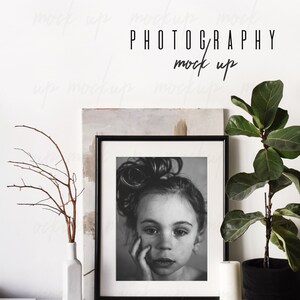 May include: Black and white photo of a young girl with her hair in a messy bun, framed in a black frame and sitting on a white shelf with a white vase, a small ball of twine, and a white candle.
