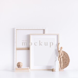 May include: Two wooden picture frames with white mounts, one slightly larger than the other, are leaning against a white wall. The larger frame has the word "mockup" printed on it. A wooden figurine, a wicker basket, and a white vase are on a white surface in front of the frames.