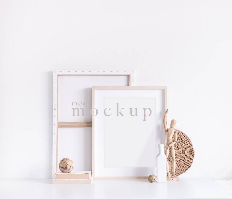 May include: Two wooden picture frames with white mounts, one slightly larger than the other, are leaning against a white wall. The larger frame has the word "mockup" printed on it. A wooden figurine, a wicker basket, and a white vase are on a white surface in front of the frames.