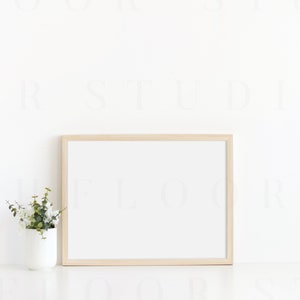 Puede incluir: Un marco de fotos de madera clara con un paspart&uacute; blanco y un fondo blanco. Una peque&ntilde;a planta en maceta con hojas verdes est&aacute; en primer plano.