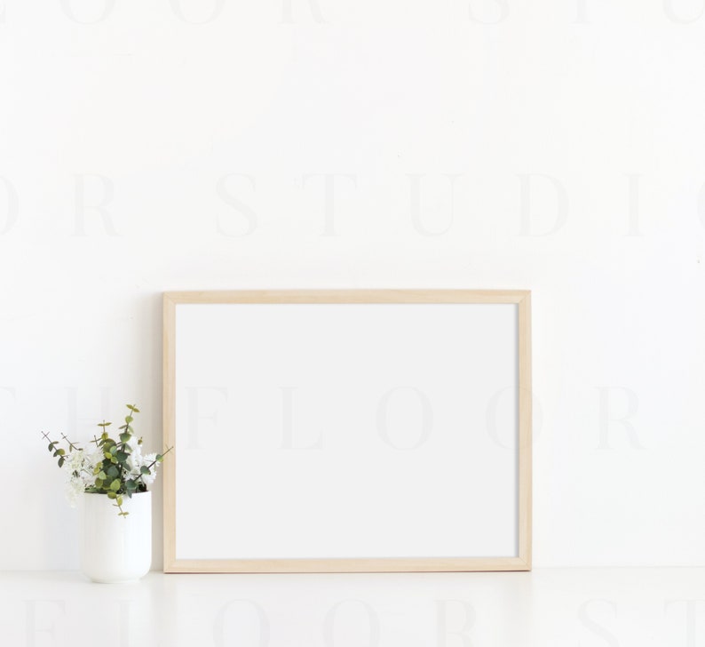 Puede incluir: Un marco de fotos de madera clara con un paspart&uacute; blanco y un fondo blanco. Una peque&ntilde;a planta en maceta con hojas verdes est&aacute; en primer plano.