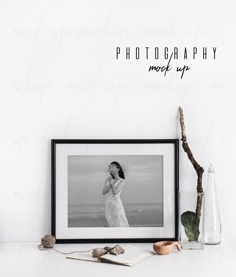 May include: Black and white photo of a woman in a white dress holding a flower in front of a beach scene. The photo is framed in a black frame and is sitting on a white table with a book, a wooden bowl, and a vase with a branch and a leaf.