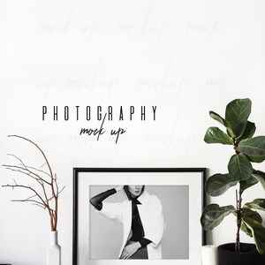 May include: Black and white photo of a person wearing a white jacket and black shirt in a black frame on a white shelf with a white vase and a potted plant. The text "PHOTOGRAPHY mock up" is written in black on the white wall behind the frame.