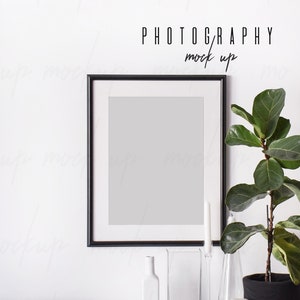 May include: Black framed photo mockup with a white mount, a potted plant, and decorative glass vases on a white surface. The text "PHOTOGRAPHY mock up" is above the frame.