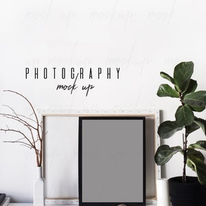 May include: A black picture frame with a white mat is leaning against a white canvas frame. A small potted plant with green leaves is on the right side of the image. The text "PHOTOGRAPHY mock up" is written in black on a white wall.