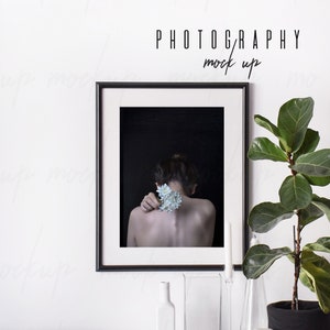 May include: A black framed photograph of a person with a flower behind their shoulder, hanging on a white wall above a table with a plant, candles, and books. The text "PHOTOGRAPHY mock up" is above the frame.