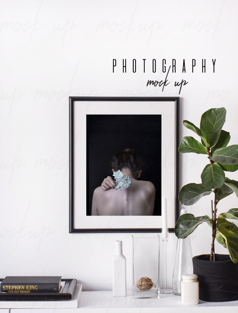 May include: A black framed photograph of a person with a flower behind their shoulder, hanging on a white wall above a table with a plant, candles, and books. The text "PHOTOGRAPHY mock up" is above the frame.