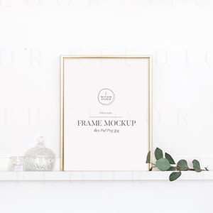May include: A gold frame mockup with a white background. The frame is sitting on a white shelf with a small glass jar and a decorative glass jar on the left side of the frame. A sprig of green leaves is on the right side of the frame.