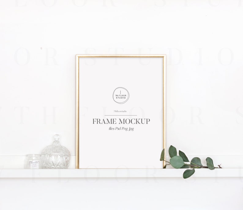 May include: A gold frame mockup with a white background. The frame is sitting on a white shelf with a small glass jar and a decorative glass jar on the left side of the frame. A sprig of green leaves is on the right side of the frame.