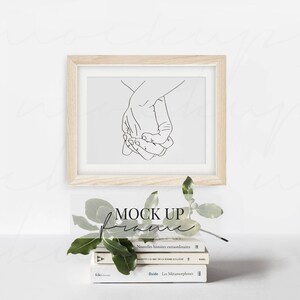 May include: A wooden frame with a white mat and a black line drawing of two hands holding each other. The frame is sitting on a stack of books with a green plant in front of it. The text "MOCK UP" is written in black on a white background.