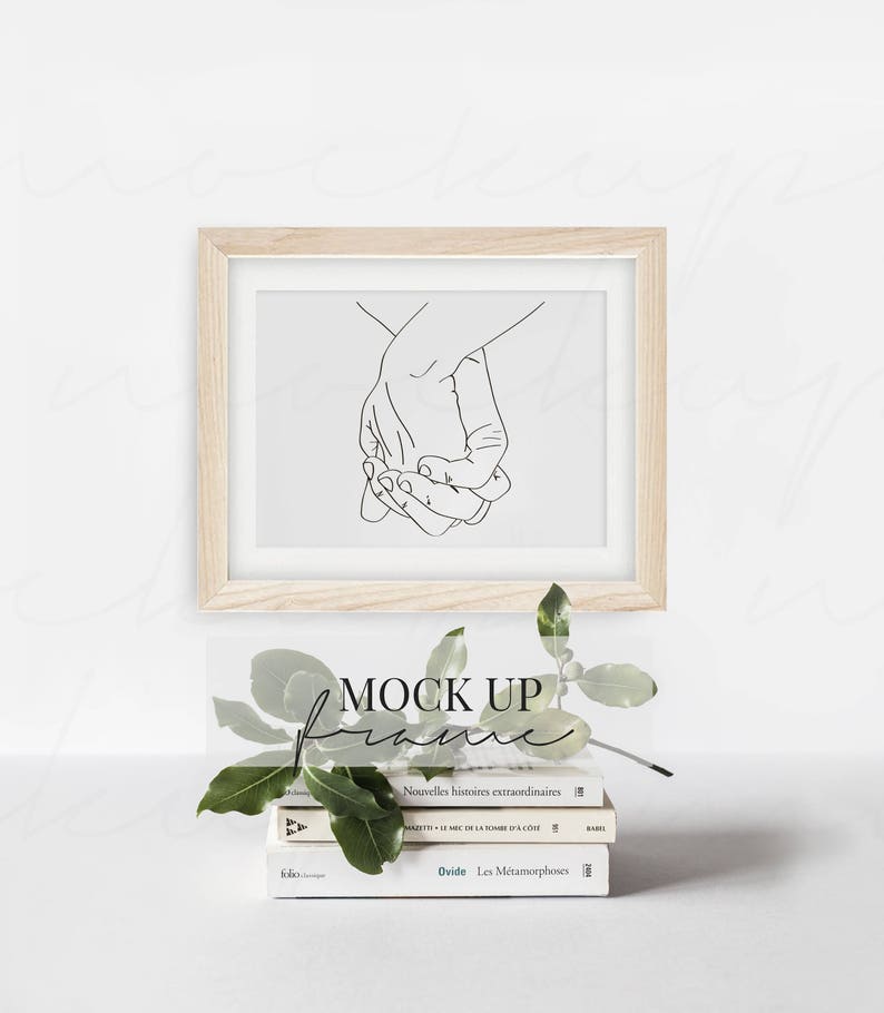 May include: A wooden frame with a white mat and a black line drawing of two hands holding each other. The frame is sitting on a stack of books with a green plant in front of it. The text "MOCK UP" is written in black on a white background.
