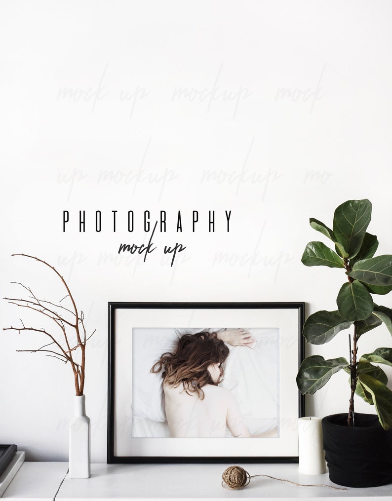 May include: Black framed photo of a woman lying on a white bed with a white wall behind it. A white vase with a brown branch is on the left side of the photo. A potted plant is on the right side of the photo. The text "PHOTOGRAPHY mock up" is written in black on the white wall.