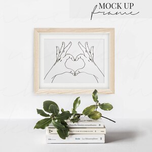 May include: A wooden frame with a white mat and a black line drawing of two hands making a heart shape. The frame is on a white wall above a stack of three books and a green plant.