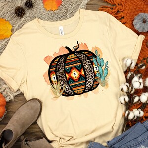 May include: A beige t-shirt with a graphic of a pumpkin with a geometric pattern and leopard print. The pumpkin is surrounded by cacti and watercolor-style leaves.
