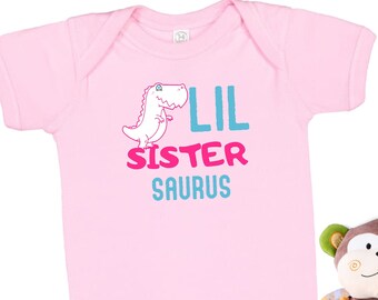Op de afbeelding: Roze baby-romper met een dinosaurus-print en de tekst "Lil Sister Saurus". De romper wordt geleverd met roze babyschoentjes en een bruin knuffel-aapje.