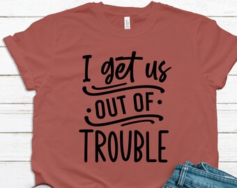 May include: A rust-colored t-shirt with black text that reads "I get us out of trouble". The shirt is laying on a white wooden surface with a pair of white sneakers and blue jeans.