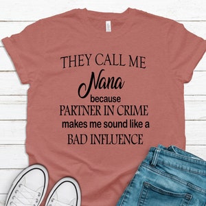 Puede incluir: Camiseta rosa polvoriento con el texto negro: "THEY CALL ME Nana because PARTNER IN CRIME makes me sound like a BAD INFLUENCE". La camiseta se combina con vaqueros azules y zapatillas blancas.