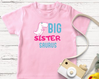 Op de afbeelding: Roze baby t-shirt met een cartoon dinosaurus en de tekst "BIG SISTER SAURUS" in blauwe en roze letters.