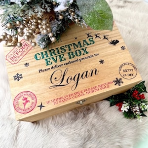 May include: A rectangular wooden Christmas Eve box with a hinged lid. The top features printed text including CHRISTMAS EVE BOX, the name Logan, and North Pole postal stamps. Decorative black snowflakes and a reindeer silhouette are printed on the light wood surface.