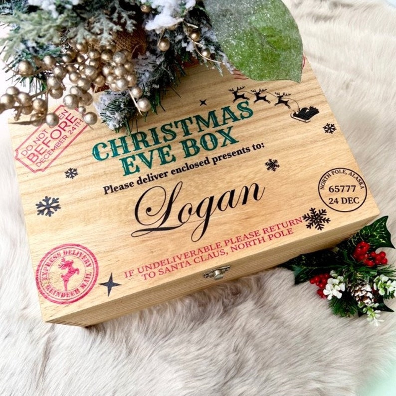 May include: A rectangular wooden Christmas Eve box with a hinged lid. The top features printed text including CHRISTMAS EVE BOX, the name Logan, and North Pole postal stamps. Decorative black snowflakes and a reindeer silhouette are printed on the light wood surface.