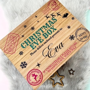 May include: A rectangular wooden box with a light brown finish, featuring the text "CHRISTMAS EVE BOX" in green, the name "Eva" in black script, and various red and black holiday-themed stamps, snowflakes, and reindeer silhouettes.