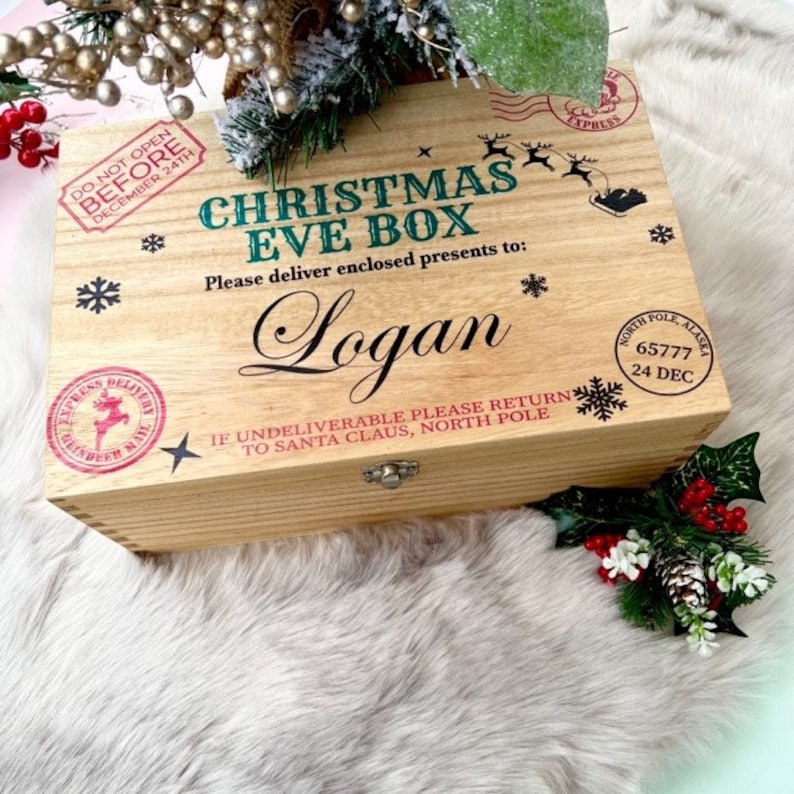 May include: A rectangular wooden box with a hinged lid featuring the text CHRISTMAS EVE BOX, the name Logan, and North Pole postal markings. The box is decorated with black snowflake icons, reindeer silhouettes, and red circular stamps.