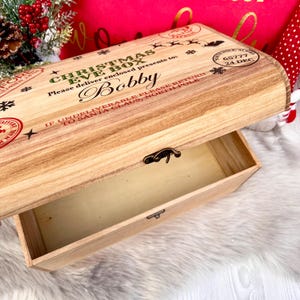 May include: A light-colored wooden Christmas Eve box with a hinged lid and metal latch. The lid features printed text including 'CHRISTMAS EVE BOX', 'Bobby', and 'NORTH POLE, ALASKA 65777 24 DEC', along with snowflake and reindeer graphics.