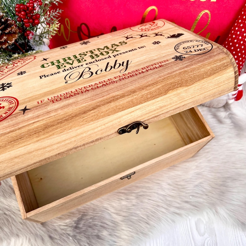 May include: A light-colored wooden Christmas Eve box with a hinged lid and metal latch. The lid features printed text including 'CHRISTMAS EVE BOX', 'Bobby', and 'NORTH POLE, ALASKA 65777 24 DEC', along with snowflake and reindeer graphics.