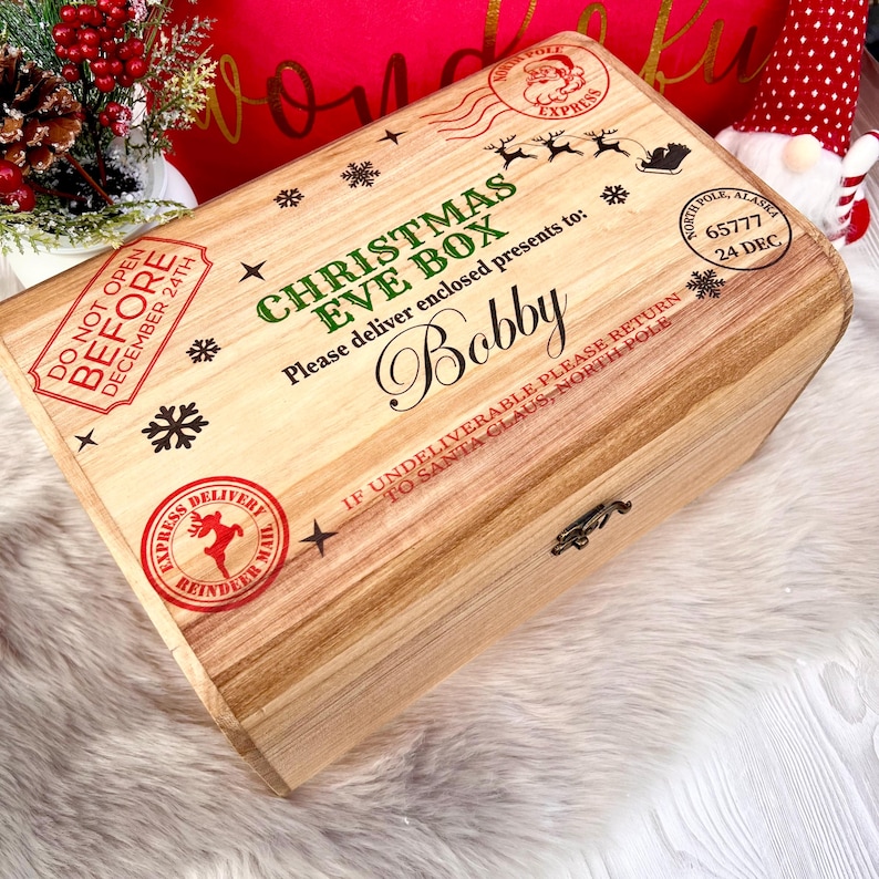 May include: A rectangular wooden Christmas Eve box featuring printed graphics of snowflakes, reindeer, and postal stamps. The text reads: CHRISTMAS EVE BOX, Please deliver enclosed presents to: Bobby, DO NOT OPEN BEFORE DECEMBER 24TH, and IF UNDELIVERABLE PLEASE RETURN TO SANTA CLAUS, NORTH POLE.