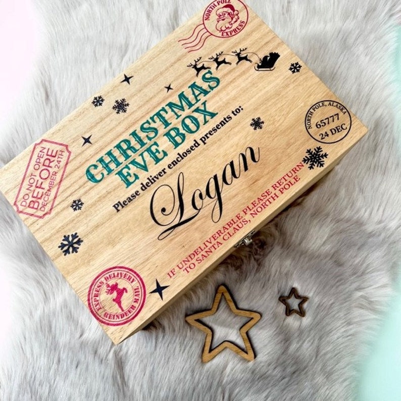 May include: A rectangular wooden box with a hinged lid featuring the text Christmas Eve Box and the name Logan in black script. The lid is decorated with red and black stamps, snowflake icons, a reindeer silhouette, and North Pole postal markings.