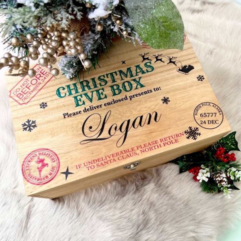 May include: A rectangular wooden Christmas Eve box with a hinged lid. The top features printed text including CHRISTMAS EVE BOX, the name Logan, and North Pole postal stamps. Decorative black snowflakes and a reindeer silhouette are printed on the light wood surface.