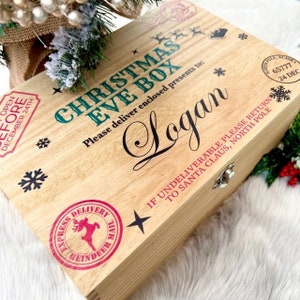 May include: A rectangular wooden box with a hinged lid featuring the text CHRISTMAS EVE BOX, the name Logan, and North Pole postal markings. The box includes decorative black snowflakes, a red reindeer stamp, and a metal latch closure.